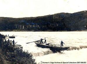 One boat traveling down river rapids. The boat is being steered by men with large oars, [Alberta].