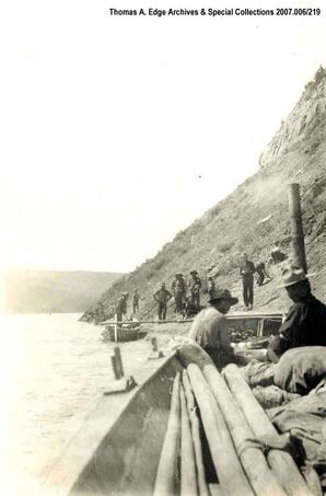Two boats landed on a shoreline. There are men in one the boats along with logs. Numerous men are standing on the shore, [Alberta]. Note on the back reads: ""Taking a rest towing scows up Athabasca River, 1910"".