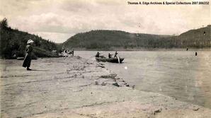 Men in a scow either landing or taking off of shore and one woman on shore watching, [Alberta]. Note on the back reads: "Mt Rapid and Portage".