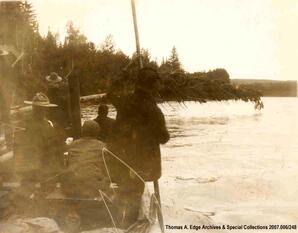 Boat full of men beside a tree which has fallen over the water. There is a rope tied to the fallen tree. This angle of the image is from downstream, [Alberta].