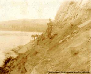 Several men standing on a steep rocky shore. There are boats in the distance traveling down a body of water, [Alberta].