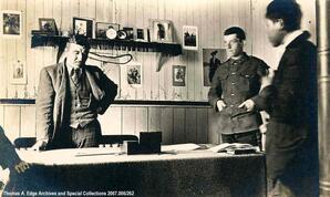 Two men and one Indian boy standing in an office. One man is behind a desk and there are numerous photographs on the wall behind him and several books on the desk, [Alberta].
