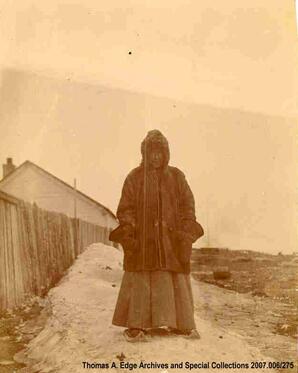 Portrait of one woman beside a fences and one building. She is dressed in a skirt and a long winter coat and she is wearing snowshoes, [Alberta]. Note on the back reads: "Dressed for the winter that has just about gone."