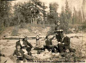 A group of seven men, woman and children sitting on a shoreline eating, [Sandy Bay].