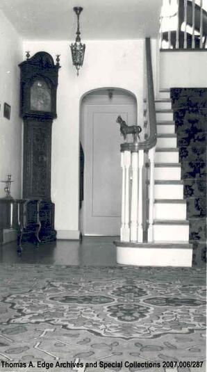 A hallway in a home with a grandfather clock, carpets, an archway and an ornate staircase, Vancouver. Note on the back reads: ""Edith Fields Home in Vancouver The old clock is here still keeping good time.""