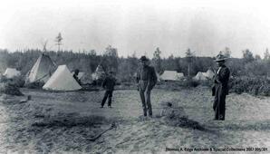 Three Indians men standing in a field of dirt, there are numerous teepees and white tents behind them, [Alberta].