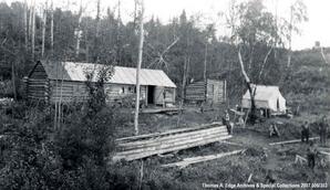 Two wooden structures and one white tent in a cleared area. There are several men working and a pile of felled logs, [Alberta].