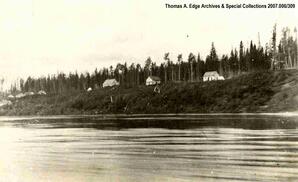 A shoreline with numerous wooden and white wooden buildings on the shore up a small embankment, [Alberta].