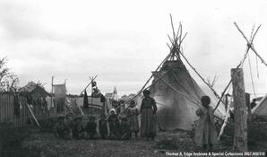 A portrait of one Indian woman and eight Indian children. They are standing within an Indian village with teepees, a wooden fence and one wooden structure, [Alberta].