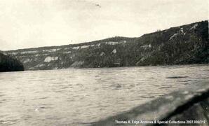 A view of a shoreline from a boat. The shoreline is tree lined, rocky and steep, [Alberta].