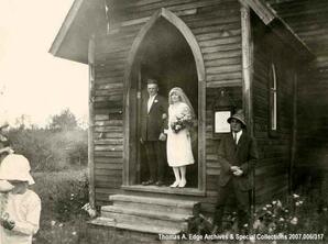 A portrait of a bride and a groom standing in an open church door. He is wearing a suit and she a white wedding gown with a veil. She is holding a bouquet of flowers, Gibbons. Note on the back reads: ""Just a wedding at Gibbons Church"".