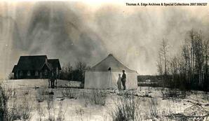 A large wooden church in a clearing with one white tent beside it. It is wintertime, Alberta].