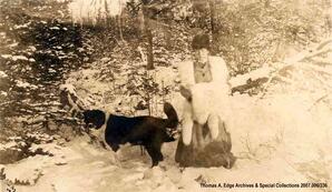 A portrait of one woman outside in the forest with her dog in winter. She is wearing a dress and a fur, Fort Chippewyan. Note on the back reads: ""Sister Edith all dolled up in Arctic White Fox, and good old Dog Rover 1910 at Chippewyan"".