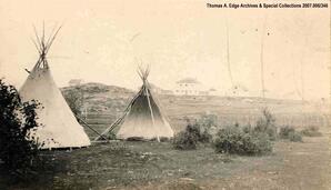 Two teepees in a field with houses in background, [Alberta]. Note on the back reads: ""Tepee at back of Fort"".