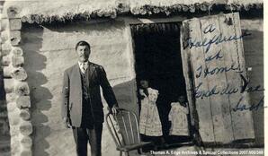 One man standing outside a house leaning on a chair. Two children are standing in the open doorway, Fond du Lac. Note on the front reads: "A Halfbreed at Home; Fond du Lac, Sask".
