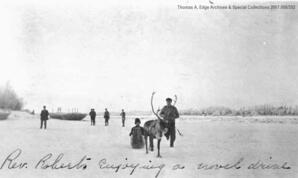 One man sitting in a sled being towed by a reindeer. There are men in the background and it is wintertime, [Alberta]. Note on the front reads: "Rev. Roberts enjoying a novel drive".