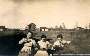 Three women eating a meal outside. Buildings are visible in the background, [Alberta].