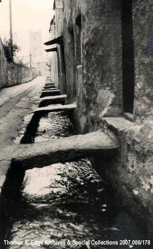 Street in Kingsteignton. Street and buildings made of stone with small stone bridge passing over creek to buildings, [United Kingdom].