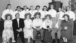 Seated group of people with some Latvian traditional clothing, and others wearing woven traditonal wreaths of Oak, Linden and Ivy leaves as well as wild flowers