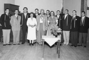 Group of men and women standing (with white dress shirts, narrow neck ties and floral headpieces)