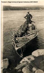 Two men in a boat, one is seated and the other is at the back holdings the oar. The boat is tied to the shore with rope, [Alberta].